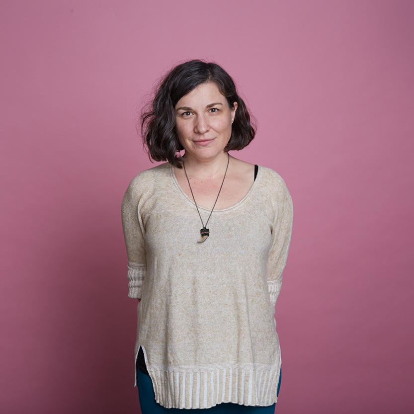 A person centred in the frame looks directly at the camera. They are wearing a beige sweater and the image is cut off at their thighs. They are on a pink background.