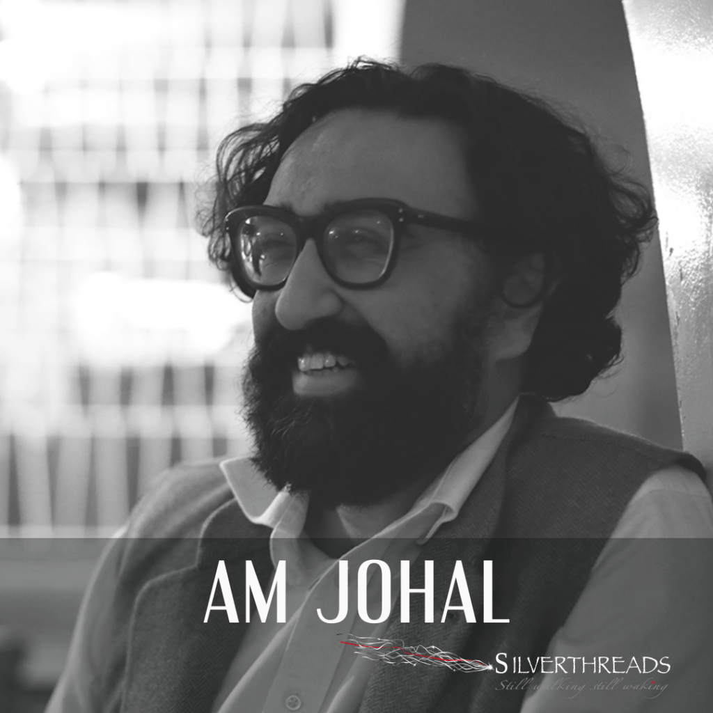 Black and white photo of Am Johal, from the shoulders up. He is sitting, leaning up against a wall, looking off to the right and smiling. He has on thick framed glasses with a beard, and is wearing a white button-down shirt with a vest.