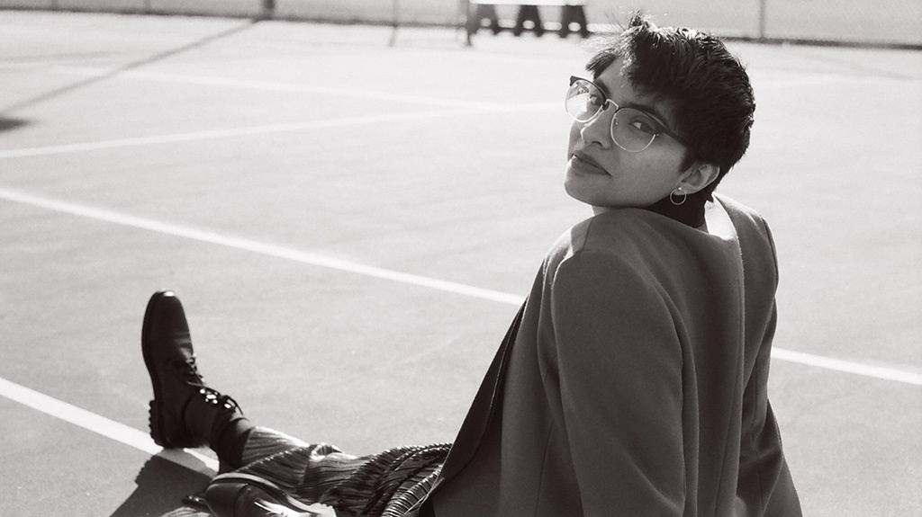 Black and white photo of Oceann, siting on a tennis court. They are wearing a blazer and looking over their shoulder at the camera.