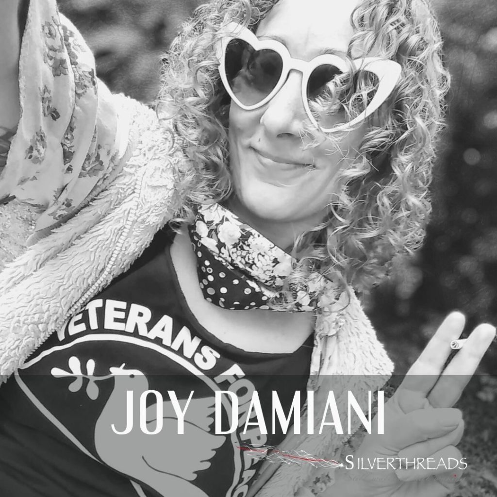 Black and White photo of Joy Damiani waist up, taking up most of the photo, smiling. She has shoulder length curly hair, wearing heart shaped sunglasses, wearing a black shirt that says in white “Veterans For Peace” with a dove on it. She has a cardigan over top of tha with a scarf tied around her neck. She is doing a kind of a peace sign with her left hand which is holding a cigarette between her fingers.