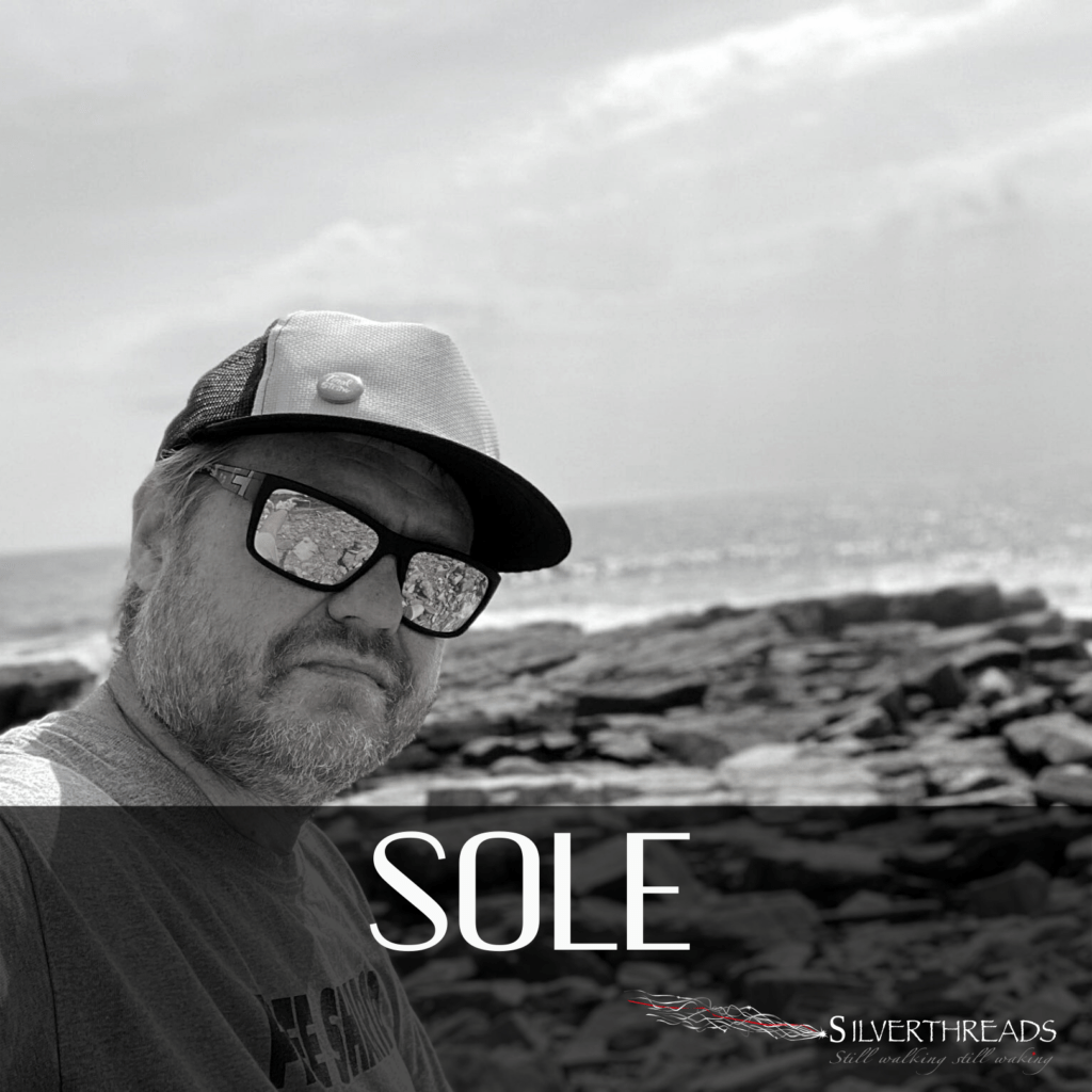 Black and white photo of Sole by the sea, on the rocks. Sole is wearing reflecting sunglasses and a snapback hat, looking unsmiling into the camera. There is a logo for Silver Threads in the bottom right corner. There is text that says “Sole”.