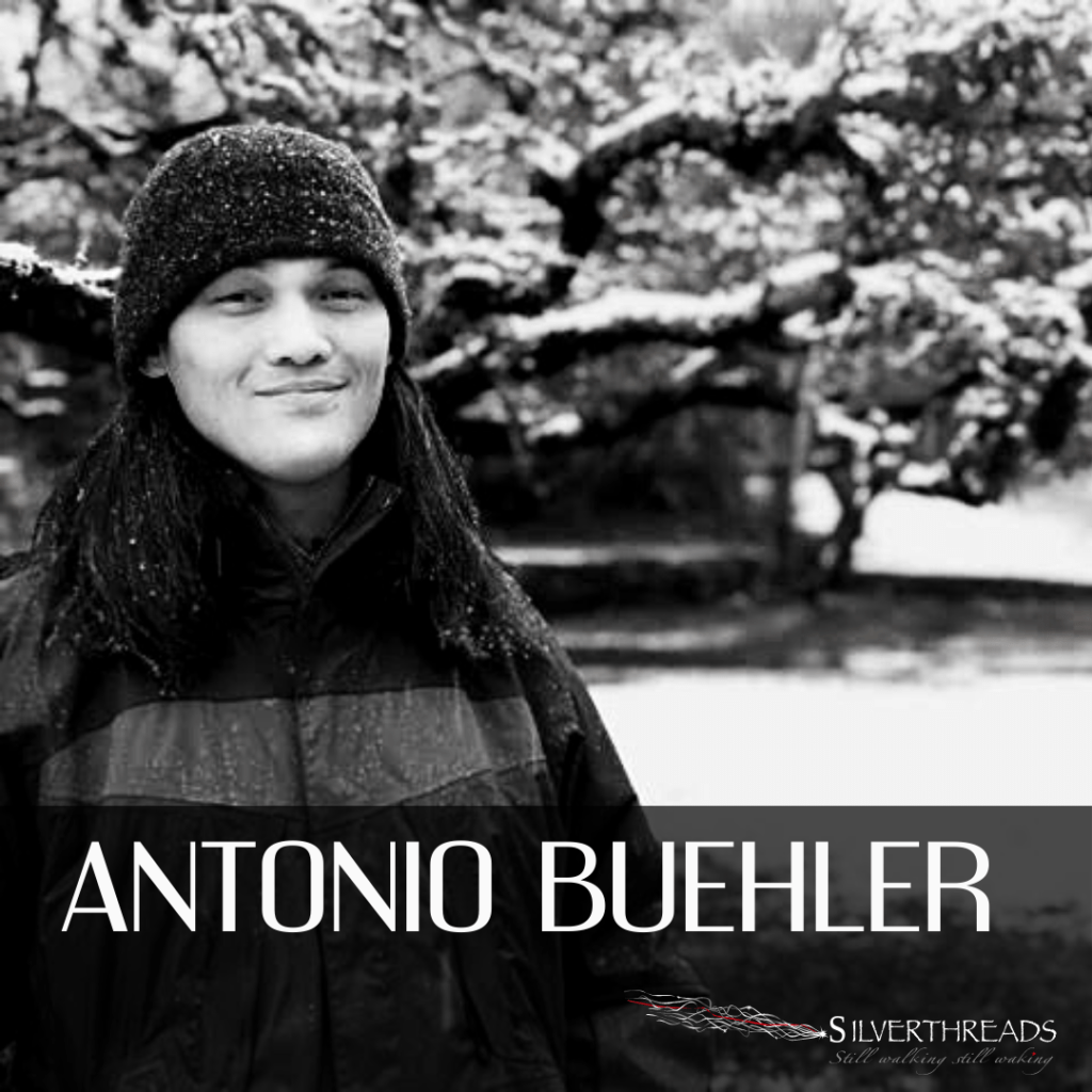 Black and white photo of Antonio. They look right at the camera and are wearing a toque and winter jacket. There is a tree covered in snow in the background. "Antonio Buehler" is in white text along the bottom, with the silver threads logo in the bottom right corner.