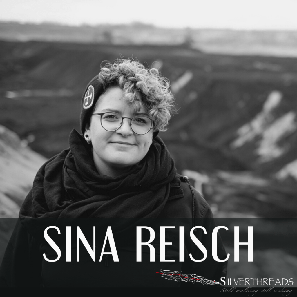 Black and white photo of Sina Reisch outside. In the distance behind her are levelled off piles of earth. She has short bleached hair, glasses, and a cozy scarf wrapped around her neck. She is smiling into the camera.