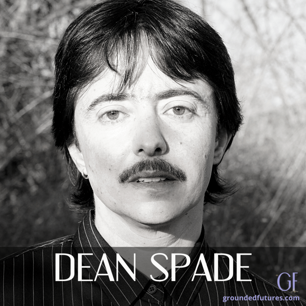A close up Black and white photo of Dean Spade. He has shortish brown hair, and a mustache and he has a serious/serene presence. He has a button up shirt on, and there are blurred trees behind him. At the bottom of the graphic, in white capital letters it says Dean Spade. To the right and below his name, in purple font is GF, and below that in purple font is says: groundedfutures.com