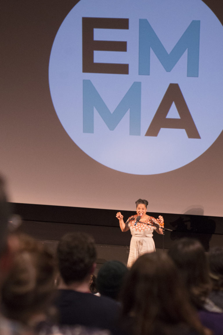 The backs of the audience, seated in a theatre, are in the foreground, with Vanessa Richards on stage below, with both hands raised up in an animated gesture. There is an EMMA Talks logo on the cinema screen behind her.