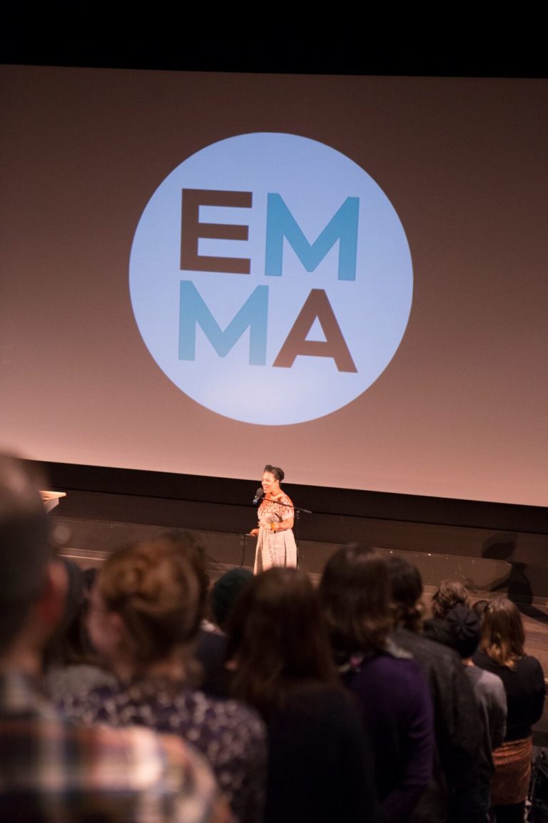 The backs of the audience, seated in a theatre, are in the foreground, with Vanessa Richards on stage below. There is an EMMA Talks logo on the cinema screen behind her.