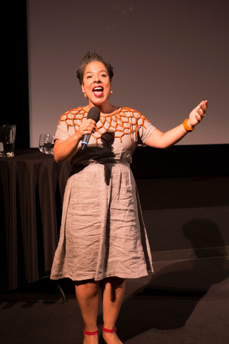 Photo of Vanessa from head-to-toe, facing towards the camera, singing into a microphone held in one hand, with the other hand raised to the crowd with palm facing upwards.