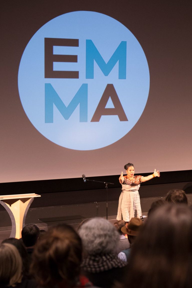 The backs of the audience, seated in a theatre, are in the foreground, with Vanessa Richards on stage below, arms open wide in front of her. There is an EMMA Talks logo on the cinema screen behind her.