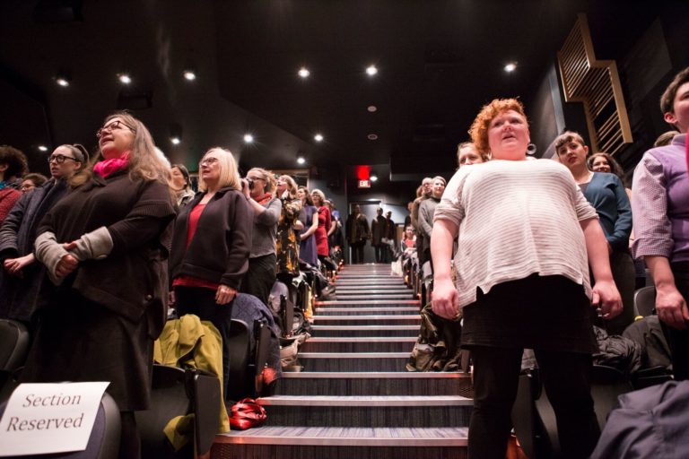 Photo with a perspective looking up the theatre stairs inside a cinema, with audience members standing in front of their seats on either side, looking forwards and singing.
