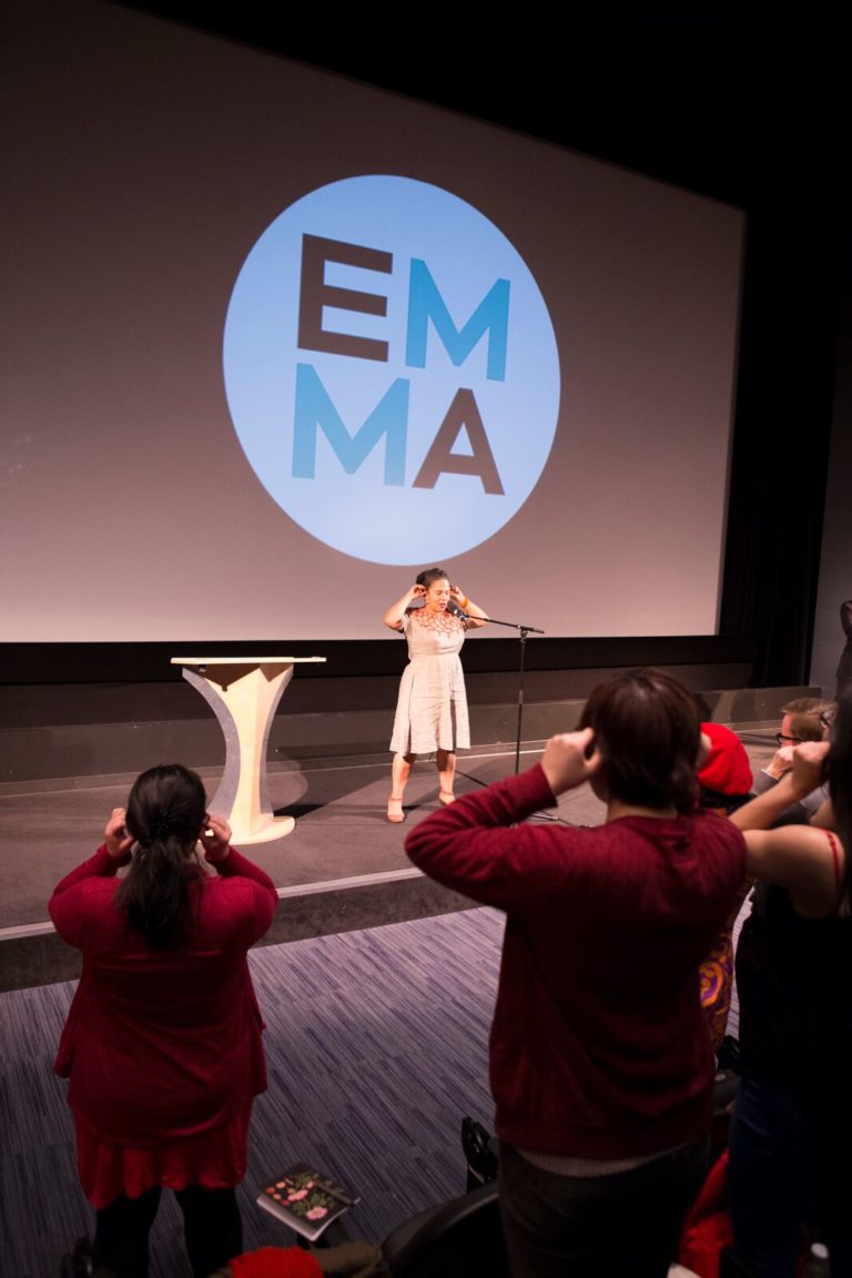 The backs of the audience, standing in a theatre, are in the foreground, with Vanessa Richards on stage below. Vanessa has both hands raised to her ears, and the crowd is mirroring her movement. There is an EMMA Talks logo on the cinema screen behind her.