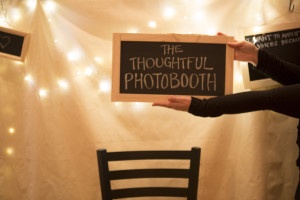 A photo of the "thoughtful photobooth" with a white backdrop, twinkle lights and a chalkboard sign held by arms extending from the right of the photograph.