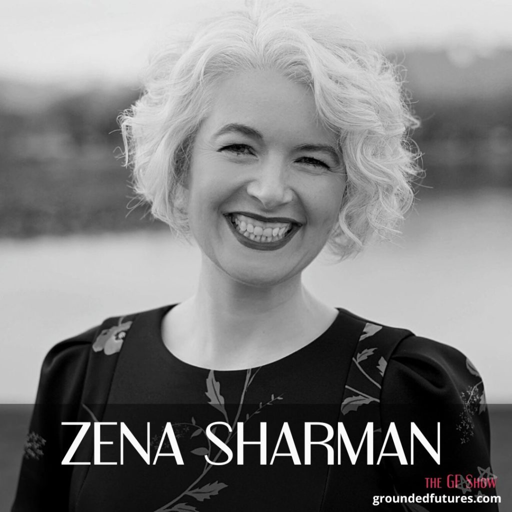 A black and white photo of Zena -- a white femme with curly, chin-length silver hair grins widely, her teeth visible. She’s wearing a black short-sleeved dress with a blue, red and green floral pattern and bold fuchsia lipstick. Her eyes are crinkled with happiness. She’s standing on a beach; the background behind her is blurred but you can make out the colours and shapes of the light brown sand, blue water and green trees behind her. There’s a landscape of snow-capped mountains in the distance.