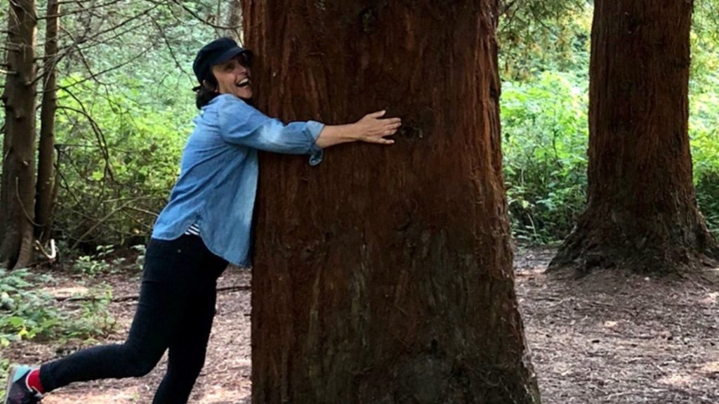 carla joyfully hugs a red cedar tree with a forest behind her.